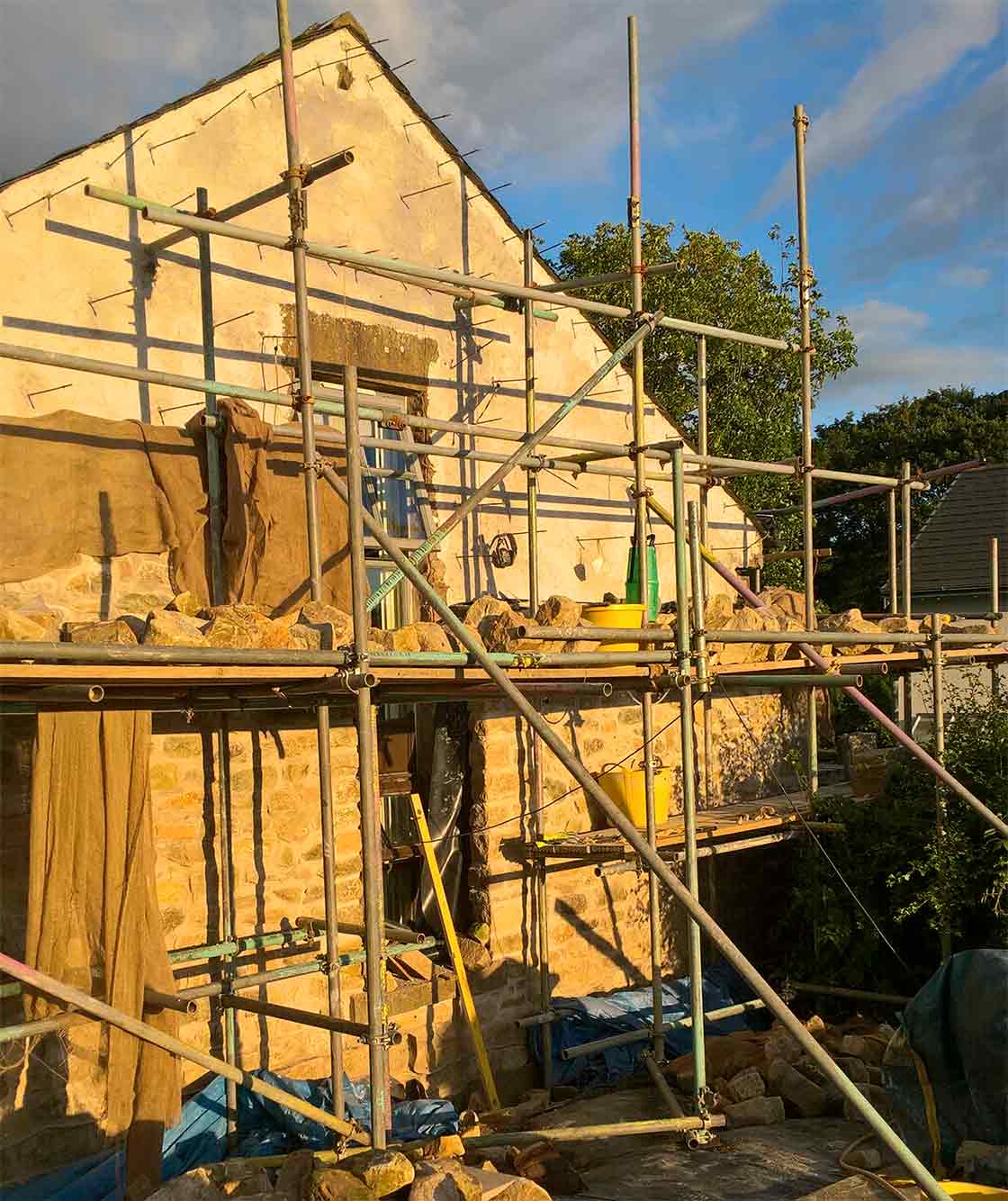 The exterior gable of Hurst’s home with a new stone skin going up to form a wide cavity for fullfill insulation, including thermally-broken Teplo-L ties and lime-plaster parge coat over the original stone, with hessian sheeting protecting fresh lime mortar.
